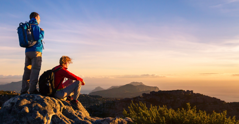 Двама души със спортни раници гледат планински пейзаж по залез