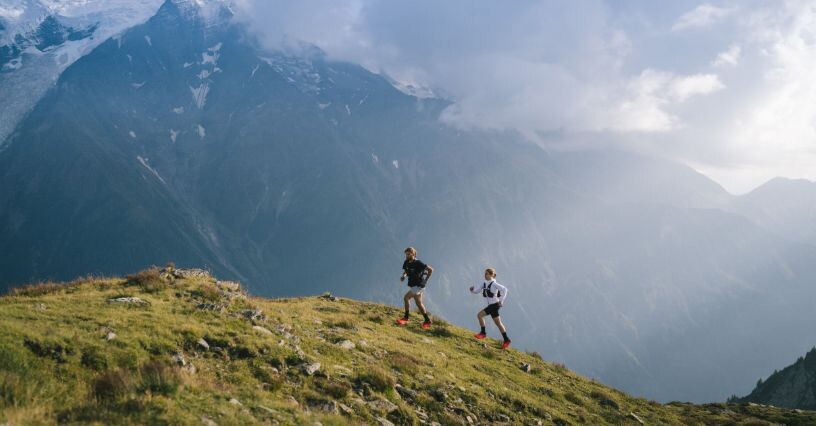 Двама души бягат по планинска пътека с изглед към впечатляващи върхове