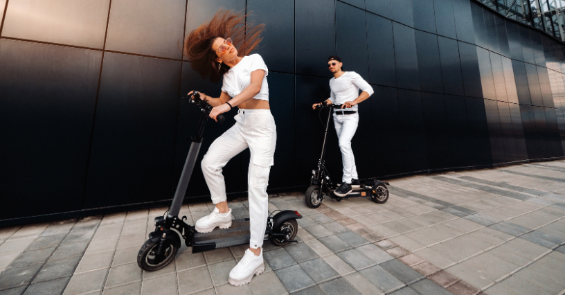Двама души се возят на електрически скутери пред модерна сграда